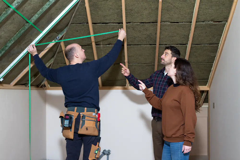 Bouwer meet met een laserafstandsmeter de dakhelling op in een zolder terwijl een stel meekijkt